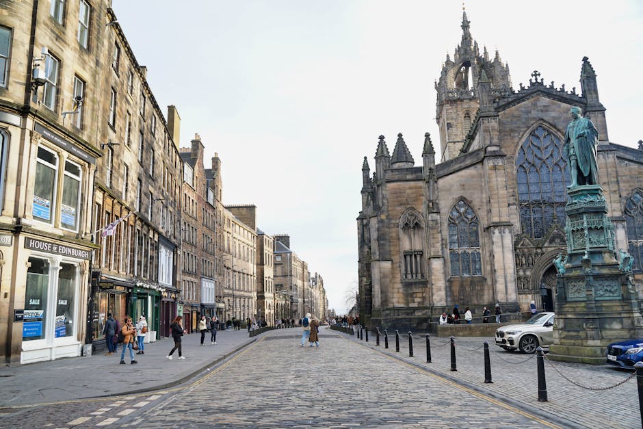 A wide view of a historic city street with cobblestone pavement and traditional-style buildings lining both sides. On the right, a large Gothic-style church with pointed arch windows, intricate stone 